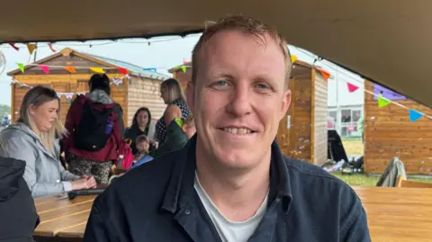 Dr Steffan Evans, smiles at the camera underneath a tent. Colourful bunting can be seen behind him. He has blue eyes and strawberry blonde short hair. 