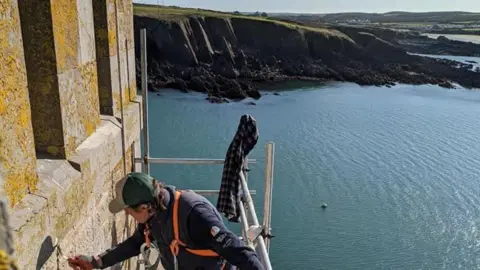 Pembrokeshire's Thorne Island fortress turned into £3m party space Mike Conner Scaffolding set up along one of the fortress walls, with a man on top of it using a hand tool. He is wearing a cap, blue gilet and orange harness. Behind the scaffolding is the sea, with cliffs in the background.