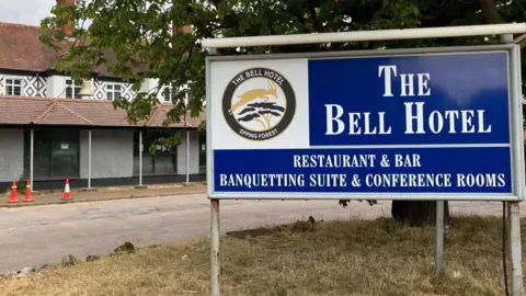 High court decision to be made on Epping asylum hotel injunction The Bell Hotel, a white building, is on the left with an entrance to the right which says The Bell Hotel, Best Western. A tree is in front of the two-storey building with three traffic cones outside.