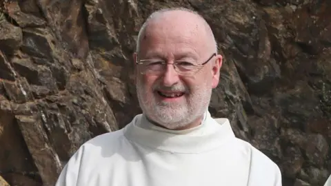 Downpatrick murder inquiry may be linked to 'brutal' attack on priest Pacemaker Fr John Murray. He is smiling at the camera. Fr Murray is bald and is wearing glasses. He has a white beard and is wearing white robes. Behind him is a rockface.