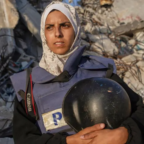 Israeli double strike on Gaza hospital AP A woman wearing a headscarf and a press tabard carried a helmet and camera strap with rubble behind her