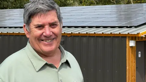 Solar-powered ice cream maker has a record sunny year in Somerset David Baker, smiling, in front of his dairy, with solar panels on the roof.