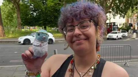 Call for free bus travel to help under-22s into work Alex Mustafa Student Alex Mustafa smiles on the street while holding a toy pigeon. She has short purple hair and is wearing lots of beaded bracelets.