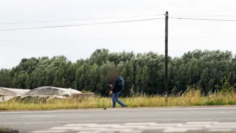 'Is my secret camera working?' Man walks along an empty road with a wood in the background.