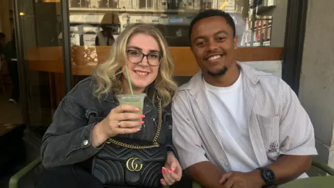 How coffee chains like Costa lost the matcha generation Lauren a blonde woman wearing a grey denim jacket, glasses with a black Gucci handbag on her lap sits next to Jordan with short black hair and a beard wearing a grey striped shirt open over a white t shirt. She is holding a green iced matcha and they're sitting outside a coffee shop.