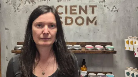 Value of small packages sent from China to UK more than doubles Katerina Buchy Katerina Buchy, a woman with long dark hair, wearing a dark grey top and a necklace, looks at the camera. The words "Ancient Wisdom" are written on the wall behind her in block capitals, and below that there is a shelf with cosmetic products on it.
