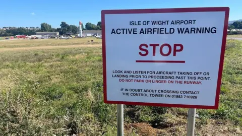 Family 'heartbroken' over fatal Isle of Wight helicopter crash A sign for Isle of Wight Airport warning of aircraft taking off or landing. A building and a stretch of grass can be seen behind the sign. It's a sunny day and the sky is blue.