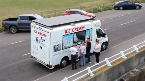 Solar-powered ice cream maker has a record sunny year in Somerset An ice cream van parked on a roadside, with four adults queueing outside. There are four solar panels attached to the van roof, covering it completely. The van has a picture menu next to a window hatch showing the ice creams it sells.