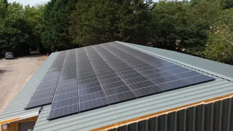 Solar-powered ice cream maker has a record sunny year in Somerset Lots of solar panels on a corrugated roof, with a car park and trees in the background.