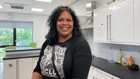 How to feed children for less in the holidays holidays Evelyn with shoulder length black hair in tight curls wearing a long sleeve black top with white floral design in a big square on the front. She is standing in a community kitchen smiling at the camera.