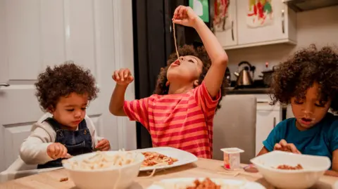 How to feed children for less in the holidays holidays Getty Images The young children sitting around a table eating spaghetti as one boy dangles spaghetti into his mouth from above his head
