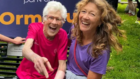 Parkrun record broken by 97-year-old Belfast runner Ormeau Parkrun Grace and Cathy are sitting on a park bench and both women are laughing. Grace is wearing a pink parkrun t-shrt and is pointing the camera. Cathy is wearing a purple t-shirt and has long hair