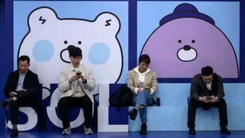 Getty Images Four men sitting in a row in a public sitting area in a metro train station and scrolling on their mobile phones in Seoul on April 8, 2025. 