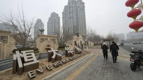 Chinese property giant to be delisted after spectacular fall AFP via Getty Images People wearing coats and hats walk past an Evergrande Group residential complex called Evergrande Palace in Beijing on 30 January, 2024 on a misty overcast day.