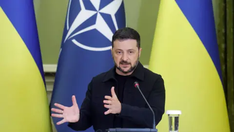 Russia trying to stop meeting on peace and prolong war, says Zelensky EPA/Shutterstock Ukrainian President Volodymyr Zelensky, dressed in his signature black, gesticulates as he addresses a joint press conference in Kyiv. Behind him his the Ukrainian flag.