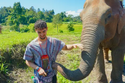 Man saw kaleidoscopic light before going blind Calum Macdonald pets an elephant as it touches his striped patterned shirt with its trunk. Behind them are green paddy fields and tropical woods beyond that. He has short black curly hair.