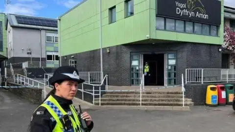 Ammanford stabbing report finds girl was fascinated by weapons and war Female police officer stands outside Ysgol Dyffryn Aman in full uniform. Police and a cordon can be seen on the steps of the green-cladded school.