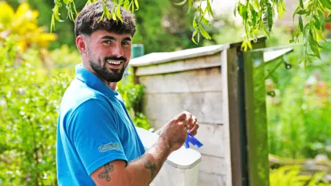 I didn't go to uni Chester Zoo Frazer Walsh in a blue Chester Zoo branded polo shirt, holding food to feed otters. He has brown hair and is smiling at the camera, in front of a glass enclosure, surrounded by trees.