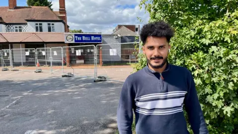 Behind the wave of asylum hotel protests Wael, a man with curly hair, is wearing a navy blue jumper. He stands in front of fences outside the Bell hotel in Epping which has temporary metal fences outside.