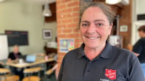 Forest of Dean school uniform pop-up a 'godsend', mum says A middle-aged woman with greying hair smiles as she stands in a Salvation Army hub. She is wearing a black Salvation Army polo shirt with a red logo. There is a woman working at a table behind her.