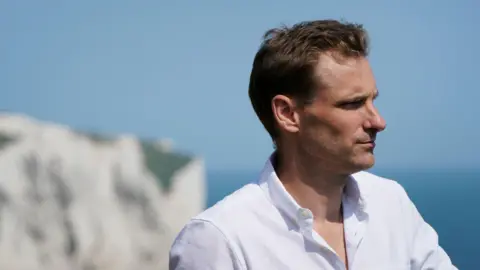 I had knife pulled on me in migrant camp, says Conservative shadow minister PA Media Chris Philp in a white shirt. The white cliffs of Dover and the English Channel are in the background
