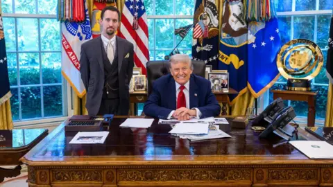 Trump's BLS nominee floats ending monthly jobs report White House Trump sits behind desk in blue suit and red tie while Anotni stands in three-piece gray suit with a beige tie, white collar and watch chain in front of windows covered in colorful flags and banners