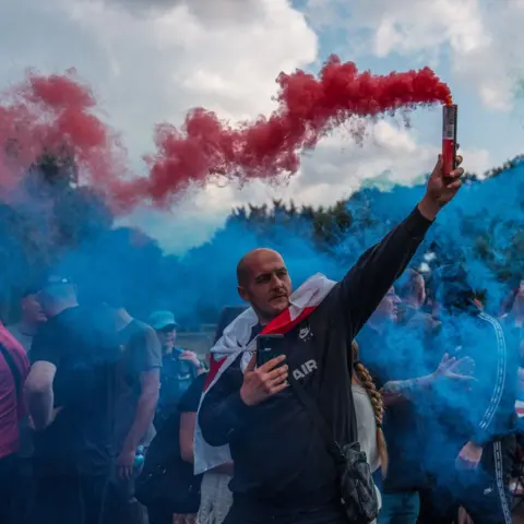 Behind the wave of asylum hotel protests Carl Court/Getty Images A man holds a flare during a protest outside The Bell Hotel on July 31, 2025 in Epping, England.