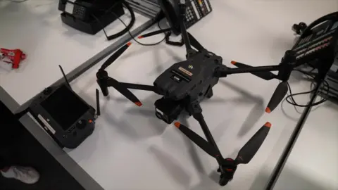 Police to deploy dedicated drone team BBC A sleek black drone with orange-tipped propellers sits on a white desk next to its remote control. The desk also has office items on it like telephones and cables.
