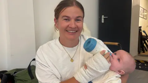BBC Ceri Jones smiles at the camera holding her baby in her left arm, holding a milk bottle. She has mousy brown hair pulled back in a ponytail and wears a white t-shirt. 