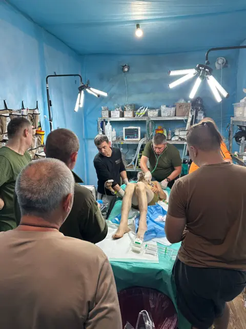 Inside Donetsk as residents flee attacks on Ukrainian region Putin wants to control A man wearing just shorts lie on a bed, surrounded by six other men. There are shelves on the walls with various medical items, and medical items also rest on the bed next to the man.
