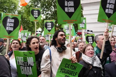 Kneecap rapper appears in court on terror charge PA Media A group of people gather holding green posters which say 'free mo chara' on them