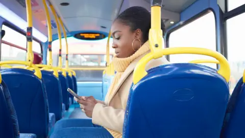 Call for free bus travel to help under-22s into work Getty Images A young woman with her hair tied back sitting sideways on a bus looking down at her smartphone