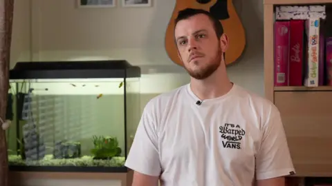 Car finance judgement 'a hard pill to swallow' BBC Marcus Johnson wears a white tee-shirt sitting in a room with a fish tank and a guitar on the wall.