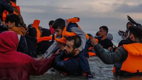 Thousands of small boat arrivals since new migrant deal with France Getty Images Migrant families wade into the sea in an attempt to board a small boat on 12 August 2025 in Gravelines, France.