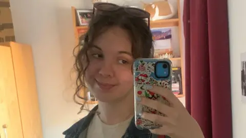 Do cash rewards or gifts boost exam grades? Jess Cooper Jess has brown curly hair tied back. She is smiling into her camera taking a selfie. There is a red curtain and brown wooden door behind her.