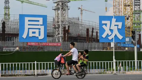Why should I care about the crisis-hit Chinese property giant? Getty Images People commute in front of the under-construction Guangzhou Evergrande football stadium in Guangzhou, China's southern Guangdong province on September 17, 2021. The photo shows a male motorcyclist with two passengers, children, seated behind him, as they ride past a large construction site.