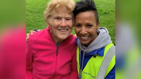 Parkrun record broken by 97-year-old Belfast runner Ormeau Parkrun Grace is wearing a pink fleece, she is sitting with Olympian Dame Kelly Holmes, Kelly is wearing a blue fleece with a yellow high-vis jacket. They are both looking at the camera and smiling