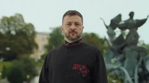 Zelensky vows to continue fighting as Ukraine marks independence day Public Broadcasting company of Ukraine A still image of President Volodomyr Zelensky, taken from a video address. Zelensky, who has short black hair and facial hair, is looking at the camera with a serious expression. He is wearing a high-necked black tunic top, which hsa a red and green pattern on the collar and the left side of the chest. He stands in front of a large, green statue and some trees, which are blurred in the background.
