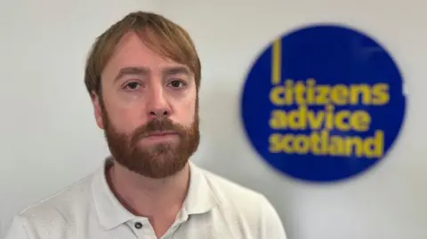 Charity says fuel poverty is year-round problem BBC David Hilferty, director of impact at Citizens Advice Scotland, sits in front of a sign displaying the charity's logo. He has brown hair and a brown beard and wears a white shirt.