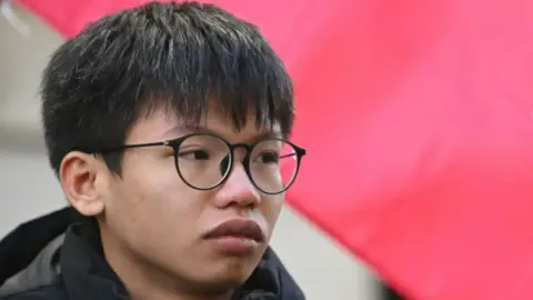 Hong Kong summons UK, Australia envoys over asylum for activists Getty Images Tony Chung, in black-rimmed glasses and a black winter jacket, attends a demonstration in London to protest the introduction of the Article 23 National Security Law in Hong Kong. Part of the Hong Kong flag is seen behind him