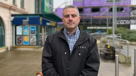 Rising Jersey food costs 'pushing families close to breadline' BBC A man, looking directly at the camera wearing a blue and white checkered top and black coat. Behind him is a building with scaffolding. The ground is pavement.