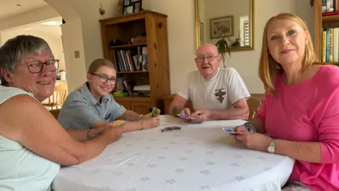 Family's multi-generation home after man's death is 'no-brainer' BBC Four people playing cards at a round table with a table cloth on. Left to right - An older woman in a light green shirt she is next to a teenage girl wearing a stripy top, next to her is a elderly bald man in a grey T-shirt and a middle aged woman with ginger hair and a pink top.