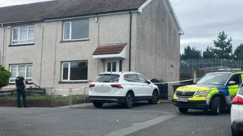 Downpatrick murder inquiry may be linked to 'brutal' attack on priest A police car sits outside a cream coloured house. There is red and white police tape cordoning off the scene.