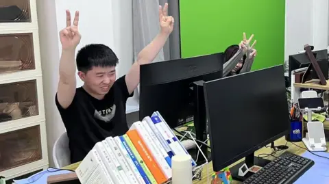 China's unemployed young adults who are pretending to have jobs BBC Shui Zhou, a young adult who pretends to have an office job, makes the victory sign with both hands that he is holding up in the air