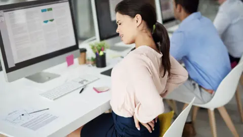 Perfect posture is a myth Getty Images Woman sitting at a chair holding her lower back and wincing in pain