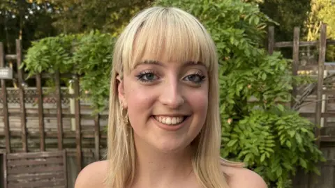 Do cash rewards or gifts boost exam grades? Imogen Farmer Imogen who has blonde mid length hair and a fringe looks at the camera smiling. Behind her is a brown garden fence and green leaves.