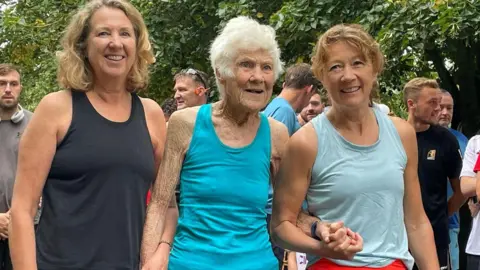 Parkrun record broken by 97-year-old Belfast runner Grace wearing a blue running top. She has white hair. To her right is a woman with blonde hair, she has a black running top. To her left is a woman wearing a grey running top, she has blonde hair. They are all holding hands.
