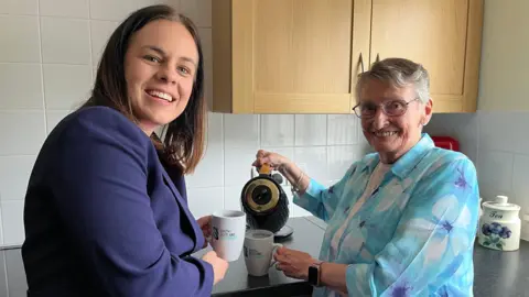 Scottish Borders social housing pilots new energy system BBC Kate Forbes, a dark-haired woman in a blue suit, holds a mug while Muriel Allison, who has grey hair and is wearing a light blue blouse, pours water from a kettle.