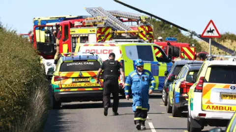 Three dead and one hurt in Isle of Wight helicopter crash Echo/Solent Emergency services including police officers and a man in all blue overalls, walking on a road with emergency service vehicles parked along it