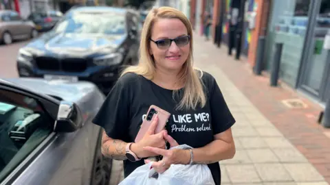School uniform costs 'extortionate' say parents in Hull Becki Bowden/BBC A woman with blonde hair and sunglasses holds her mobile phone and shopping bags as she smiles into the camera. She is standing on a street with shops on the right and parked cars on the left.